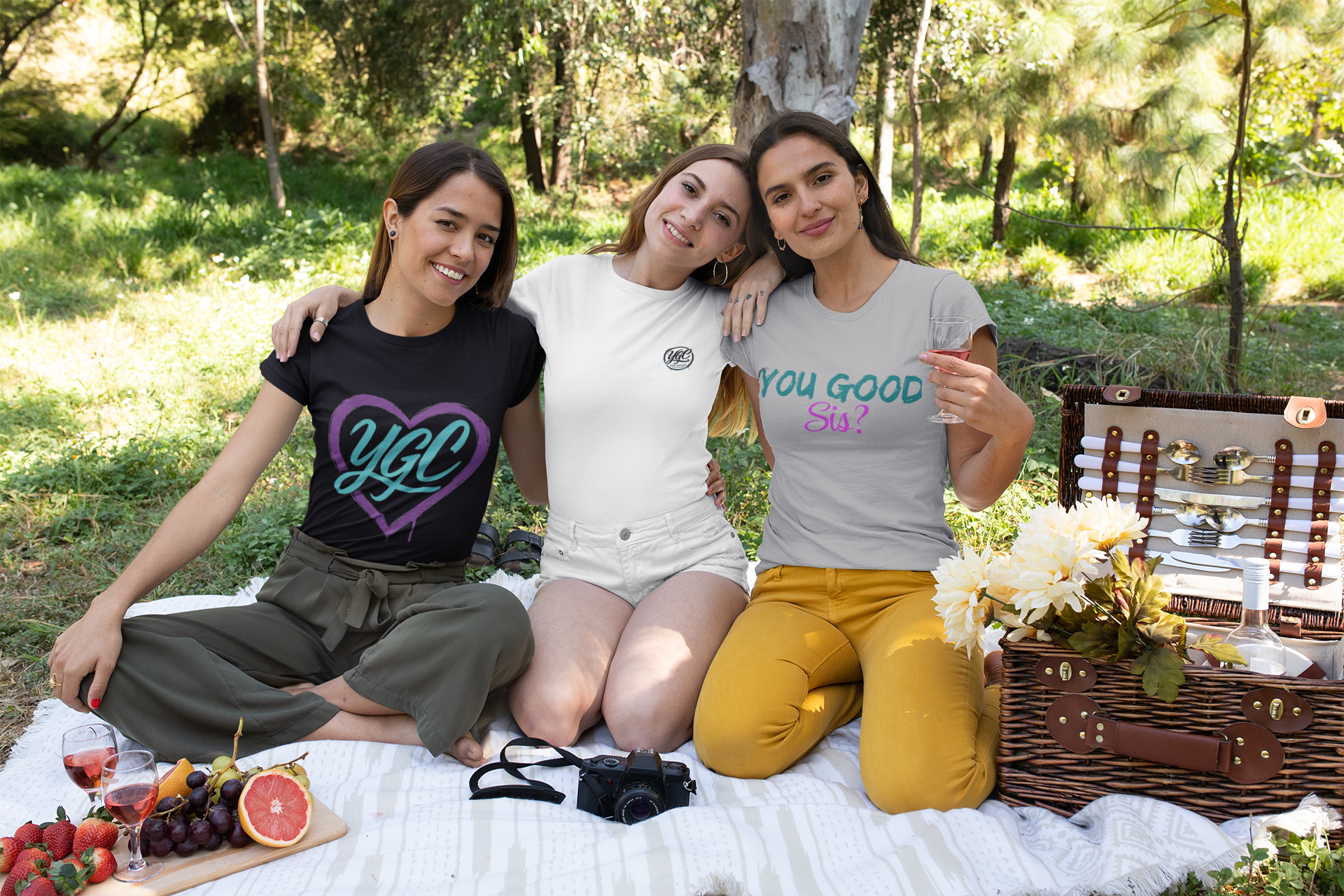 Three women sitting on a blanket in a park, wearing t-shirts with visible text.